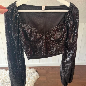 Svart glittrig croppad topp från Nelly - Säljer en svart croppad topp från Nelly med långa puffärmar och glittriga paljetter över hela tyget. Toppen har en snygg sweetheart-ringning och är perfekt för nyår. Materialet är syntet och toppen har en figurnära passform.