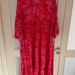 Rosa och röd blommig långklänning Cellbes - Säljer en färgstark långklänning från Cellbes med blommigt mönster i rosa och rött. Klänningen har v-ringad hals, långa ärmar och ett luftigt fall. Perfekt för dig som gillar att sticka ut med färg och mönster.