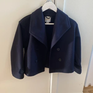 Mörkblå skepparkavaj från Gina Tricot - Stilren mörkblå skepparkavaj från Gina Tricot med bred krage och dubbelknäppning framtill. Jackan har långa ärmar med knappdetalj vid ärmslut och är tillverkad i ett mjukt ull-liknande material. Perfekt för dig som vill ha en klassisk look.