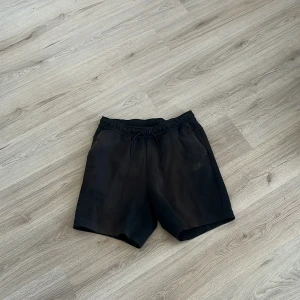Svarta Nike tech fleece shorts - Svarta shorts från Nike med elastisk midja och snörning. De har en clean look, sidofickor och en bakficka med dragkedja. Perfekta för chill dagar eller träning. Materialet känns mjukt och sportigt.