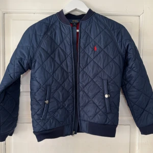 Quiltad blå jacka Polo Ralph Lauren - Säljer en quiltad mörkblå jacka från Polo Ralph Lauren med röd logga på bröstet. Jackan har dragkedja, ribbade muddar och två fickor med knapp framtill. Insidan är röd och materialet är polyester. Perfekt för höst och vår!