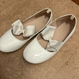 Vita ballerinaskor med rosett från H&M - Söta vita ballerinaskor från H&M med rund tå och en stor dekorativ rosett framtill. Skorna har en glansig finish och är perfekta för dig som gillar en klassisk och feminin stil.