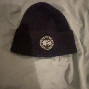 Svart mössa från Canada Goose - Svart ribbstickad mössa från Canada Goose med klassisk rund logotyp framtill. Tjockt och varmt material, perfekt för kalla dagar. Enkel och stilren design som funkar till de flesta outfits.