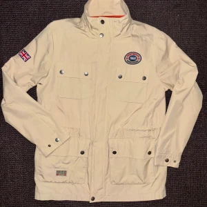 Morris jacka med huva i perfekt skick - Mycket snygg vindjacka/ tunn parkas i storkek L från Morris med broderad patch på bröstet och brittisk flagga på ärmen. Jackan har fyra fickor framtill med tryckknappar, hög krage och avtagbar huva. Perfekt för dig som gillar sportig och klassisk stil. Mycket fint skick och inga tecken på användning. Passar även dig som har storlek Medium.