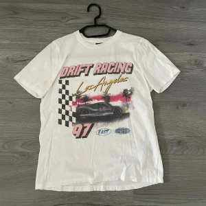 Vit t-shirt  - Säljer en vit t-shirt med coolt Drift Racing-tryck och Los Angeles-text i guld. Framsidan har motiv med racerbilar, palmer och schackrutigt mönster. T-shirten är kortärmad och har en avslappnad passform, perfekt för dig som gillar streetstyle och motorsport.