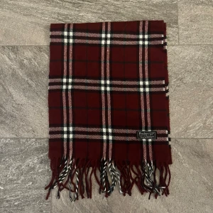 Vinröd rutig halsduk från Burberry - Klassisk vinröd halsduk från Burberry med rutigt mönster i vitt, svart och rött. Halsduken har tvinnade fransar i ändarna och är tillverkad i mjuk ull. Perfekt accessoar för att lyfta din vinterstil.