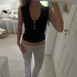 Svart ribbad  linnetopp H&M - Svart croppad linnetopp från H&M med ribbad struktur och djup v-ringning. Toppen har tre snygga guldfärgade knappar framtill och är ärmlös för en clean look. Perfekt att styla med jeans eller kjol för en trendig vibe.