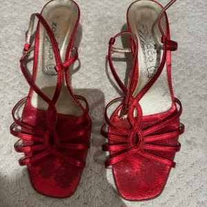 Röda metallic sandaler med klack - Snygga röda sandaler från Zodiaco med smala remmar och öppen tå. Skorna har fyrkantig tå, låg klack och justerbar rem runt vristen. 