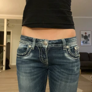 Låga Miss Me bootcut jeans med detaljer - Säljer ett par blå Miss Me jeans med bootcut och låg midja. Jeansen har tyvärr ett litet hål på högra knät som jag visar på bilden men annars är dom i bar skick☺️