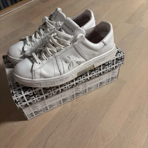 Vita sneakers från Premiata  - Snygga vita sneakers från Premiata i mjukt skinn med klassisk rund tå och snörning. Skorna har diskreta sömdetaljer, logga på sidan och blå Premiata-tryck på hälen. Perfekta för dig som gillar stilrena och trendiga sneakers. På den högra skon saknas en del så OBSERVERA det innan ni köper! 