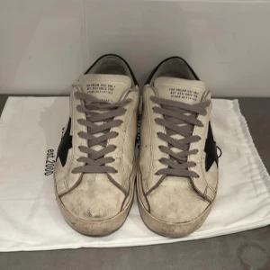 Golden Goose vita sneakers med svart stjärna - Golden Goose sneakers i vitt skinn med ikonisk svart stjärna på sidan och svart hälparti med logga. Grå snörning och beige detaljer vid tån. Platt sula och rund tå. Perfekt för dig som gillar en cool och avslappnad streetstil.