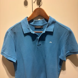 Blå pikétröja från J.Lindeberg - Snygg blå pikétröja från J.Lindeberg med klassisk krage och korta ärmar. Tröjan har en liten vit logga på bröstet och är gjord i mjuk bomull. Perfekt för en clean och sportig look.