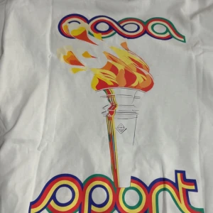 Vit Casablanca Sport t-shirt med fackla - Vit t-shirt från Casablanca Sport med färgglad tryckt fackla och logga i rött, blått, grönt och gult på bröstet. Klassisk passform och rund hals. Perfekt statement-plagg för dig som gillar sportig och retro stil. Oanvänd