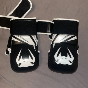 Svarta Venom boxningshandskar M - Säljer ett par svarta och vita boxningshandskar från Venom i storlek M. Handskarna har coolt grafiskt tryck med Venom-loggan och breda kardborreband för extra stöd. Materialet är syntetiskt och designen är riktigt sportig med vita detaljer och kontraster. Pris går att diskutera