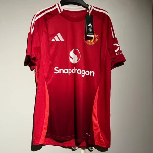 Manchester United Adidas matchtröja M - Manchester Uniteds officiella matchtröja från Adidas i rött med vita detaljer och klubbmärke broderat på bröstet. Tröjan har korta ärmar, klassiska vita ränder på axlarna och är tillverkad i lätt, ventilerande polyester.