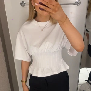 Vit korsett-topp med kort ärm - Säljer en vit korsett-topp med korta ärmar och rund hals. Toppen har en snygg, figurnära passform med markerad midja och veckade detaljer framtill. Materialet känns mjukt och lätt, perfekt för att skapa en clean och trendig look.