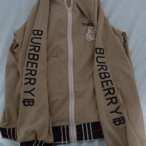 Beige Burberry bomberjacka med tryck - Snygg beige bomberjacka från Burberry med svart logotyptryck längs ärmarna och klassiskt rutigt mönster på krage och muddar. Jackan har dragkedja framtill och en liten nalle-illustration på bröstet. Perfekt för dig som vill sticka ut med en ikonisk streetstyle-look.