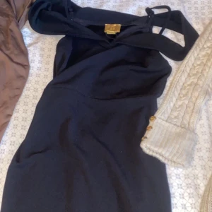 Svart axelbandslös klänning - Snygg svart klänning med axelbandslös design och stilren siluett. Klänningen har en elegant form och ett guldfärgat spänne framtill som detalj. Perfekt för dig som gillar minimalistisk och chic stil. Passar dig som vill sticka ut med en klassisk look.