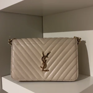 Beige quiltad axelväska  - Snygg beige axelväska från YSL med guldiga detaljer och kedja. Väskan har ett quiltat mönster i V-form och klassisk YSL-logga framtill. Tillverkad i skinn och har en stilren, rektangulär form med flera fack på insidan.