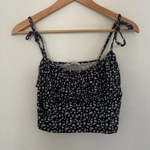 Svart blommig linnetopp från FB Sister - Supersöt svart linnetopp från New Yorker med vita fjärilar och knytbara axelband. Croppad modell med lätt rynkade detaljer framtill. Det är ett mjukt och skönt material som är stretchigt. På vänster sida vid ryggen så är axelbandet nästan av men går lätt att fixa med nål och tråd. 