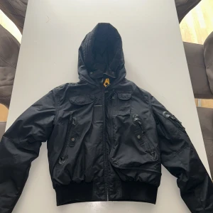 Svart Parajumpers pufferjacka med huva - Grym svart pufferjacka från Parajumpers Masterpiece Series med flera fickor, huva och coolt PJS-märke på ärmen. Jackan har dragkedja, ribbade muddar och orange detaljer på insidan. Perfekt för kalla dagar och riktigt snygg streetstil.