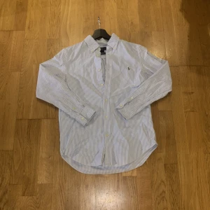 Randig skjorta från Ralph Lauren - Klassisk skjorta från Ralph Lauren i vitt och blått med tunna ränder. Skjortan har lång ärm, knappar framtill och broderad logga på bröstet. Tillverkad i bomull och har en tidlös, normal passform. Storlek 16 men tycker den passar runt 170 som bäst