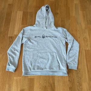 Grå hoodie från Sail Racing - Snygg ljusgrå hoodie från Sail Racing med svart logga på bröstet. Tröjan har huva, magficka och långa ärmar. Mjuk insida i bomullsblandning, perfekt för kyliga dagar. Enkel och stilren design som funkar till det mesta.