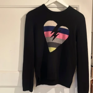 Svart stickad tröja Zadig & Voltaire - Svart stickad tröja från Zadig & Voltaire med ett stort hjärta i flera färger och en blixt på bröstet. Tröjan har rund halsringning och långa ärmar. Perfekt för dig som vill ha en cool statement-piece i garderoben.