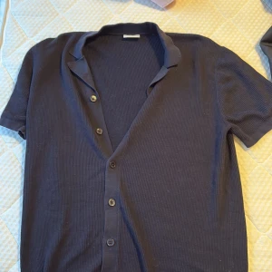 Mörkblå skjorta  - Snygg mörkblå skjorta med korta ärmar och ribbad struktur. Skjortan har klassisk krage och knäppning framtill med svarta knappar. Perfekt för dig som gillar enkel och stilren design med lite edge.