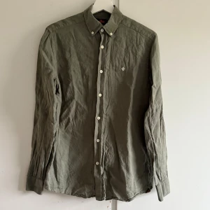 Olivgrön linneskjorta från Morris XS - Olivgrön skjorta från Morris i 100% linne. Klassisk modell med button-down krage, vita knappar och broderad logga på bröstet. Perfekt för dig som gillar en stilren och avslappnad look. Lite urtvättad 