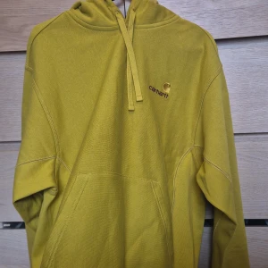 Gul hoodie från Carhartt S - Säljer en snygg hoodie från Carhartt i färgen golden olive. Tröjan har broderad logga på bröstet, huva med snörning och stor känguruficka framtill. Perfekt för dig som gillar streetwear och vill sticka ut med en cool gul färg.