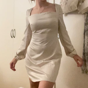 Satin klänning - Luscious satin dress i storlek 34/XS från Nelly. Krämfärgad/beige i färgen med snörning baktill. I mycket bra skick då den tyvärr aldrig kommit till användning. 
