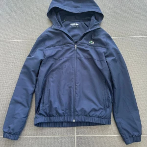 Lacoste Sport vindjacka blå - Snygg marinblå vindjacka från Lacoste Sport med huva och dragkedja framtill. Jackan har elastiska muddar, två fickor och klassisk krokodillogga på bröstet. Tillverkad i 100% polyester med meshfoder för extra komfort. Perfekt för dig som gillar sportig och clean stil. Har dock ett litet hål vid armen men ska laga det!