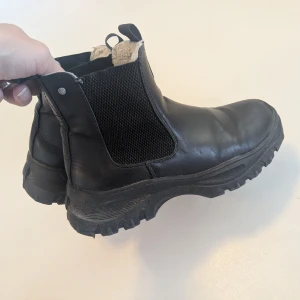 Svarta varmfodrade Chelsea boots - Säljer ett par svarta Chelsea boots med grov sula och elastiska paneler på sidorna. Skorna är fodrade med mjukt foder för extra värme och har dragflikar både fram och bak. Tillverkade i skinn med robust känsla, perfekta för kyligare dagar. Lite skavda fram på tårna men i övrigt bra skick
