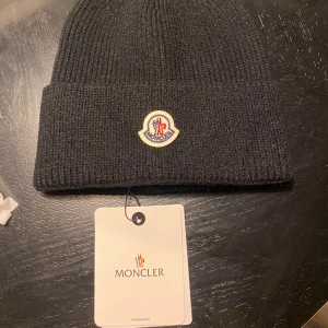 Svart och vit Moncler mössa i ull - Två ribbstickade mössor från Moncler, en svart och en vit. Båda har uppvikt kant och Monclers klassiska broderade logotyp framtill. Tillverkade i mjuk ull som håller dig varm och snygg under kalla dagar. Perfekt accessoar för vinter och höst.
