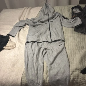 Grå Nike tech set byxor + tröja 500kr - Snygg ljusgrå hoodie från Nike med hel dragkedja och huva. Tröjan har svarta detaljer, Nike-logga på bröstet och en ficka på ärmen. Perfekt för chill eller träning, tillverkad i mjuk bomullsblandning.