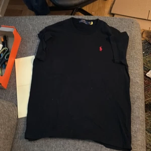 Svart t-shirt från Polo Ralph Lauren - Svart t-shirt från Polo Ralph Lauren med klassisk rund hals och röd broderad logga på bröstet. T-shirten är kortärmad och tillverkad i mjuk bomull, perfekt för en clean och stilren look.