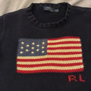 Mörkblå stickad tröja Polo Ralph Lauren - Snygg mörkblå stickad tröja från Polo Ralph Lauren med amerikansk flagga i rött, vitt och blått på bröstet samt RL-broderi. Tröjan har rund hals och långa ärmar med breda ribbade muddar. Perfekt för dig som gillar klassisk preppy stil.