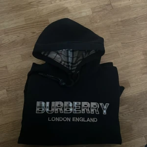 Svart Burberry hoodie med logga - Svart hoodie från Burberry med stor logga framtill och klassiskt rutigt foder i huvan. Tröjan har snörning vid huvan och långa ärmar. Perfekt för dig som vill ha en stilren och lyxig streetwear-look.