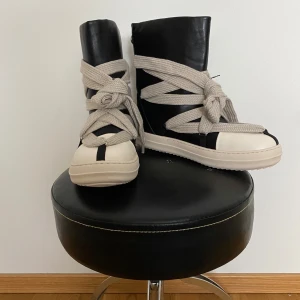 Rick Owens DRKSHDW high-top sneakers - Unika boots i svart och vitt med breda, beige snören som går omlott runt hela skon. Snygg kontrast mellan den svarta skaftdelen och den vita, rundade tån. Platt sula och högt skaft ger en cool siluett. Tillverkade i skinn för en lyxig känsla.