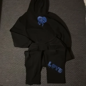 Svart hoodie med blått LOVE-tryck - Svart hoodie med huva och blått graffiti-inspirerat LOVE-tryck på bröstet och ryggen. Texten 'L'amour guérit' under trycket på ryggen. Hoodien har långa ärmar och är i mjukt material, perfekt för chill dagar.