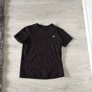Svart t-shirt från Lyle & Scott - Svart t-shirt från Lyle & Scott med klassisk passform och rund hals. T-shirten har ett litet broderat gult logomärke på bröstet och är tillverkad i mjuk bomull. Perfekt för en clean och stilren look.