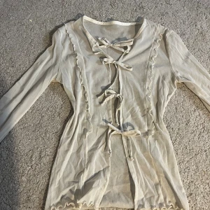 Beige långärmad blus med knyt - Supersöt beige blus med långa ärmar och flera knyt framtill. Toppens material är tunt och lätt, med volangdetaljer längs kanterna och vågig finish vid ärmslut och nederkant. Perfekt för dig som gillar romantiska och trendiga detaljer.