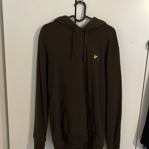 Mörkbrun hoodie från Lyle & Scott L - Säljer en mörkbrun hoodie från Lyle & Scott i storlek L. Tröjan har huva med snörning, känguruficka och den klassiska gula fågel-loggan broderad på bröstet. Perfekt för chill dagar och enkel att matcha med jeans eller joggers.