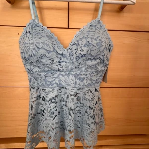 Ljusblå spetslinne med volang - Supersöt ljusblå linnetopp med tunna axelband och v-ringning. Toppen är helt täckt av blommig spets och har en volangkant nedtill som ger en romantisk vibe. Perfekt att styla med jeans eller kjol för en fräsch look.