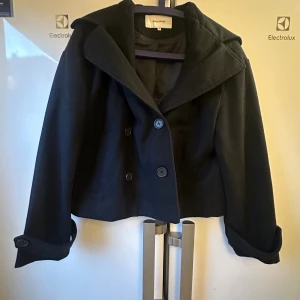 Bubbleroom, short coat.  - Stilren mörkblå kort kappa  från Bubbleroom  med bred krage och dubbelknäppning framtill. Jackan har långa ärmar med knappdetalj vid ärmslut och är tillverkad i ett mjukt ullmaterial. Perfekt för dig som gillar en klassisk men modern look.