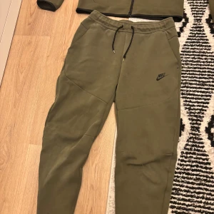 Olivgrön Nike tech dress - Nike Tech Fleece dress i olivgrönt med hel dragkedja och huva. Snygg svart Nike-logga på bröstet och diskret svart detalj på ärmen. Litet hål på ena knät