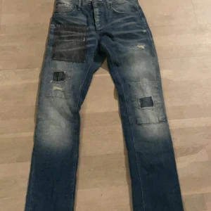 Patchade blå jeans från Jack & Jones - Säljer ett par blå jeans från Jack & Jones med coola patchade detaljer och slitningar på benen. Jeansen har raka ben, klassisk femficksdesign och tydliga kontrastsömmar. Materialet är jeans i bomull och de har en normal passform.