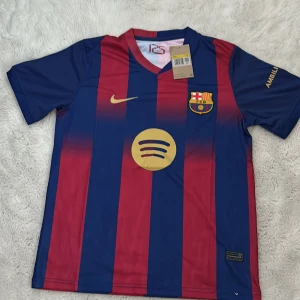 Barça matchtröja Nike S - Barcelona fotbollströja från Nike i storlek S. Tröjan har klassiska blå och röda vertikala ränder, guldiga detaljer som Nike-logga och Spotify-logga på bröstet samt klubbmärket. Tillverkad i lätt polyester med korta ärmar och V-ringad krage.