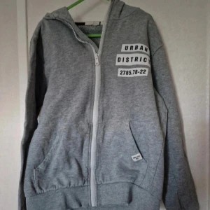 Grå hoodie med tryck - Grå hoodie med dragkedja från Kappahl. Stora vita tryck med svart text både fram och bak, där det står bland annat 'Urban District' och flera ord på ryggen. Trycket på bröstet har det blivit lite sprickor i. Tröjan har huva, fickor och ribbade muddar. Perfekt för en streetinspirerad look.